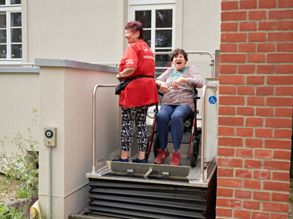 Zwei Frauen nutzen einen Lift, eine Frau sitzt auf einem Rollator.